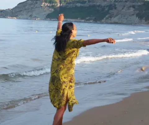 Dunja tanzt am Strand, im Hintergrund das Meer und Berge.